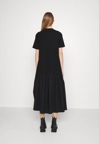 Schwarzes Kleid mit kurzen Ärmeln, lockerer Passform und gerafftem Saum. Hergestellt aus weichem Stoff, mit knöchellangem Design und kombiniert mit schwarzen Ankle-Boots.