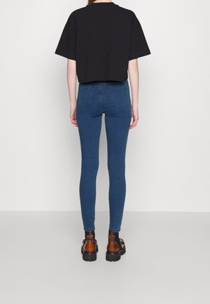 Osoba stojąca tyłem, ubrana w obcisłe, wysokiej talii niebieskie dżinsy, czarny krótki T-shirt oraz brązowe masywne buty, na jednolitej szarej podłodze.