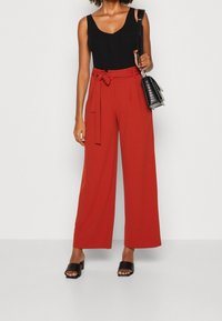 Pantalons larges rouges avec une ceinture à nouer, associés à un haut sans manches noir et des sandales noires à brides. Un simple sac à main noir comme accessoire.
