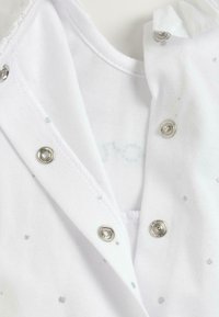 Vêtement blanc avec motif de points argentés, présentant des fermetures à pression en métal et une texture douce. Col visible et bords soigneusement cousus.