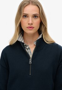 Pull-over à col zippé bleu marine en maille texturée, superposé à une chemise à col rayée bleu clair et blanche. Fermeture éclair avec quincaillerie en argent.