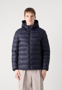Giacca puffer blu navy con cappuccio, caratterizzata da sezioni trapuntate, chiusura frontale con cerniera e un piccolo etichetta con il marchio sul petto.