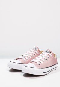 Růžové plátěné tenisky s nízkým profilem, bílé gumové podrážky, černý pruh a klasický design na šněrování. Obsahují kovové očky a štítek s logem.