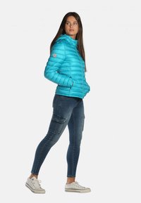 Doudoune bleue avec capuche, présentant un design matelassé et un écusson de logo sur la manche. Associée à un jean cargo ajusté et des baskets.