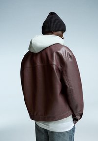 Veste en cuir bordeaux avec un sweat à capuche gris en dessous, associé à un jean bleu clair. Un bonnet en tricot noir couvre la tête.