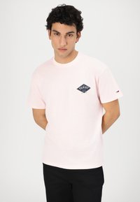 Camiseta de algodón rosa claro con mangas cortas, que presenta un logo de diamante azul en el pecho y un pequeño detalle de logo en la manga.