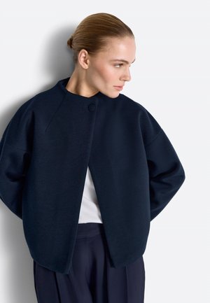 Ženska z gladko nazaj počeščenimi lasmi, oblečena v temno mornarsko modro veliko jakno čez belo majico, stoji pred belim neutemeljenim ozadjem in gleda desno.