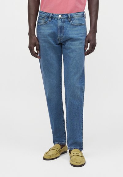 Homme portant un jean bleu droit, des mocassins jaunes et une chemise rose, debout devant un fond uni.