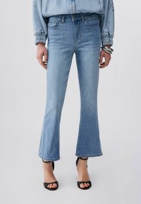 Femme portant un jean flare court bleu clair, une veste en denim assortie, des talons ouverts noirs et des bracelets argentés au poignet.