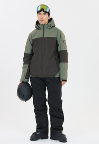 Skijacke mit einem farblich abgetrennten Design in Olivgrün und Dunkelbraun, kombiniert mit schwarzen Skihosen und einem schwarzen Helm. Skibrille auf der Stirn.
