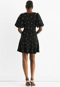 Black dress with puff sleeves, embellished with small white dots, flared skirt, and keyhole back detail. Worn with strappy black heels.