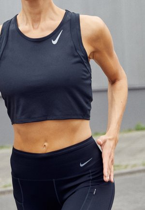 Atletyczna osoba biegająca na zewnątrz, ubrana w czarny krótki top Nike i legginsy z widocznymi logo marki na piersi i biodrze.
