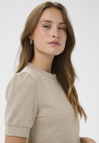 Beżowa bluzka z krótkim rękawem, okrągłym dekoltem i bufkowymi detalami na ramionach, wykonana z miękkiego materiału, o subtelnej fakturze i gładkim wykończeniu.