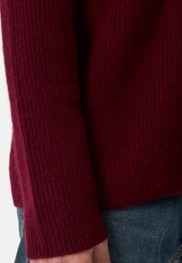 Burgundy-ribbad tröja med en mjuk textur, har en avslappnad passform och breda ärmslut. Kombinerad med blå denimjeans.