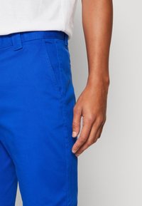 Klart blå bomullsbukser med rak framkant, sidofickor och en mjuk textur, tillsammans med en vit skjorta som syns ovanför.