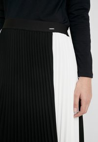 Plissé rok met een brede zwarte tailleband, met aan de ene kant zwart en aan de andere kant wit, wat de contrasterende kleuren en textuur benadrukt.