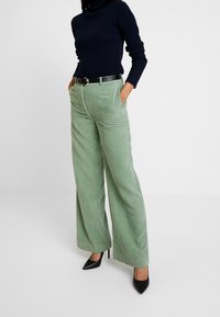 Kobieta w jasnobeżowych szerokich spodniach z korkowej tkaniny, czarnym pasku, granatowym swetrze z golfem w prążki oraz czarnych, spiczastych szpilkach, stoi z rękami w kieszeniach.