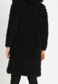 Sort faux pelsfrakke med en lang, afslappet pasform, lange ærmer og en blød, tekstureret overflade. Kanten falder lige over knæene.