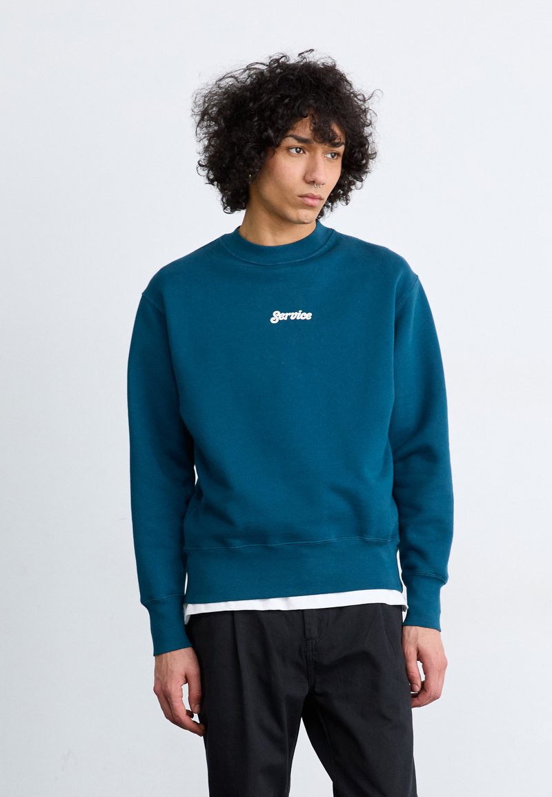Teal crewneck sweatshirt with ribbed cuffs and hem, featuring white embroidered "Service" text on the front. Worn over a white shirt.