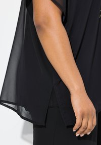 Schwarze durchsichtige Bluse mit kurzen Ärmeln, die einen seitlichen Schlitz und ein geschichtetes Design aufweist. Die glatte Textur ergänzt die fließende Silhouette.