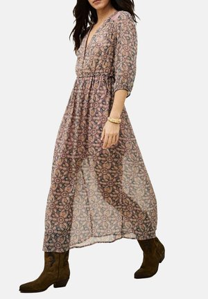 Femme portant une robe maxi à manches longues et à motifs, avec une taille cintrée, associée à des bottines en daim marron et des bracelets au poignet.