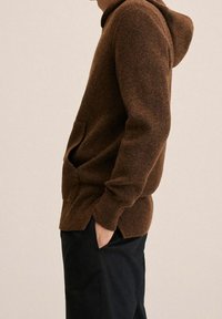 Hoodie marron en maille, confectionné en tissu texturé, doté d'une poche avant, d'un ourlet côtelé et de fentes latérales. Porté avec un pantalon foncé.