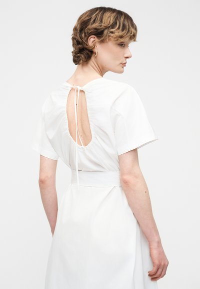 Γυναίκα με κοντά κυματιστά μαλλιά που φοράει λευκό φόρεμα με κοντά μανίκια και ανοιχτή πλάτη δεμένη με λεπτά κορδόνια.