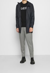 Wasserabweisende schwarze Jacke mit Kapuze, über einem schwarzen Pullover und grauen karierter Hose geschichtet, ergänzt durch schwarze Knöchelstiefel.
