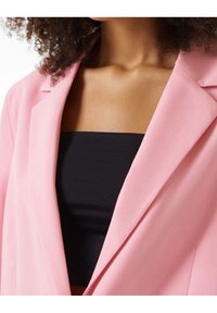 Růžová blazer s elegantním střihem, s ostrými klopami a hladkou texturou, nošená přes černý top. Zaměření na výstřih a barevný kontrast.