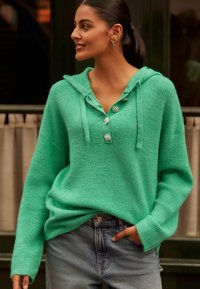 Roheline kudumihüüe, millel on struktureeritud viimistlus, esiosas nööbiga sulgemine ja nööri, koos helesinine teksapükstega.