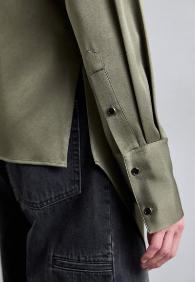 Olivgrünes Langarmhemd mit glatter Textur, ausgestattet mit einem Manschette mit schwarzen Knöpfen und einem seitlichen Schlitz, kombiniert mit dunklen Jeanshosen.