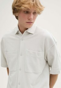 Korte mouwen, lichtgrijs shirt van een gestructureerde stof, met een knopenfront en twee opgestikte zakken op de borst.