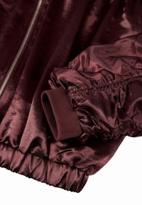 Vyno raudonos spalvos bomberio striukė, pasižyminti blizgiu, tekstūruotu audiniu, elastingais rankogaliais ir sidabrine užtrauktuku, einančiu per priekį.