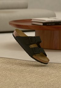 Μαύρο σανδάλι με δύο ρυθμιζόμενους ιμάντες και πάτο από φελλό που αιωρείται πάνω από υφασμένο μπεζ χαλί, ξύλινο τραπεζάκι σαλονιού και βιβλία στο παρασκήνιο.