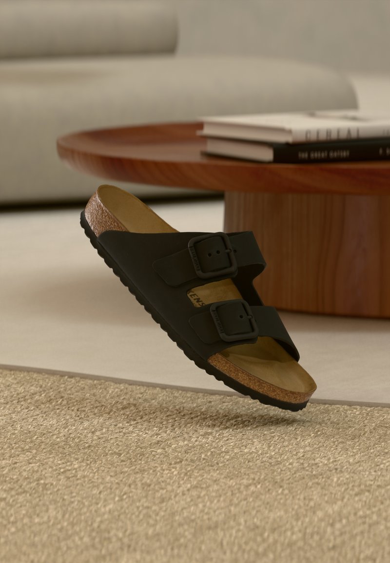 Μαύρο σανδάλι με δύο ρυθμιζόμενους ιμάντες και πάτο από φελλό που αιωρείται πάνω από υφασμένο μπεζ χαλί, ξύλινο τραπεζάκι σαλονιού και βιβλία στο παρασκήνιο.