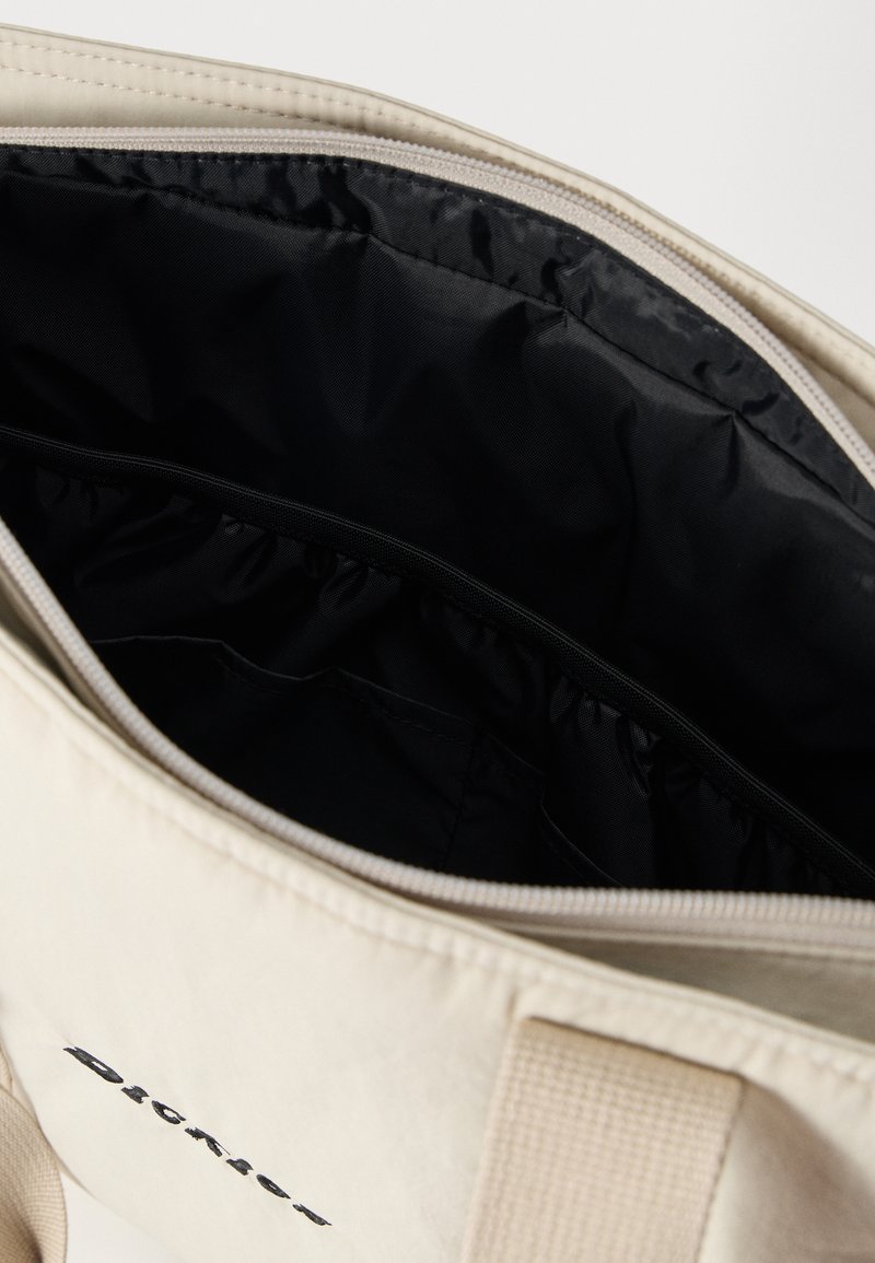 Borsa shopper beige con interno in tessuto nero, chiusura con cerniera e tasche elastiche interne per organizzare piccoli oggetti.