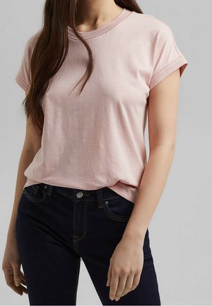 Kobieta ubrana w jasnoróżowy t-shirt z krótkim rękawem i ciemnoniebieskie dżinsy na jednolitym szarym tle.