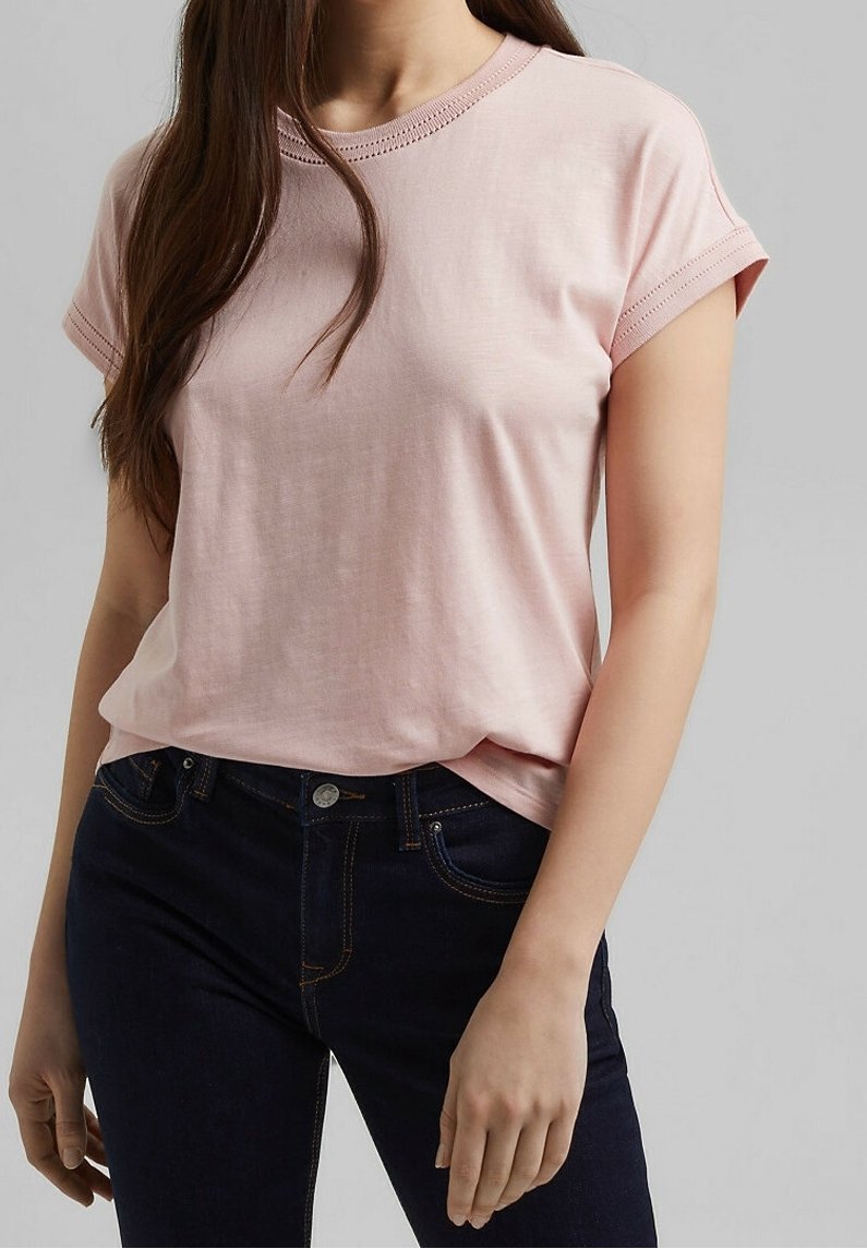 Femme portant un t-shirt rose clair à manches courtes et un jean bleu foncé sur un fond gris uni.
