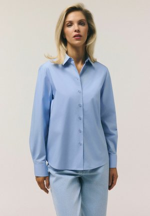 Blondynka w jasnoniebieskiej koszuli na guziki i jasnoniebieskich dżinsach, stojąca na tle jednolitego beżowego tła.