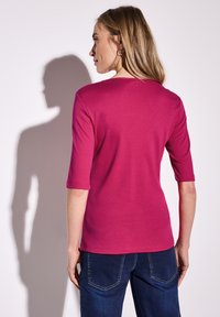 Magenta bawełniana koszulka z trzema czwarte rękawami, okrągłym dekoltem i gładką fakturą. Połączona z ciemnoniebieskimi dżinsami.