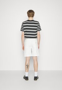 Sort- og gråstribet kortærmet skjorte, hvide shorts, sorte sko og ankelsokker; synlig slange-tattoo på venstre ben.