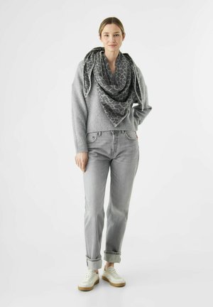 Femme portant un pull gris, une écharpe à imprimé léopard, un jean gris avec des ourlets, et des baskets blanches, se tenant devant un fond blanc.