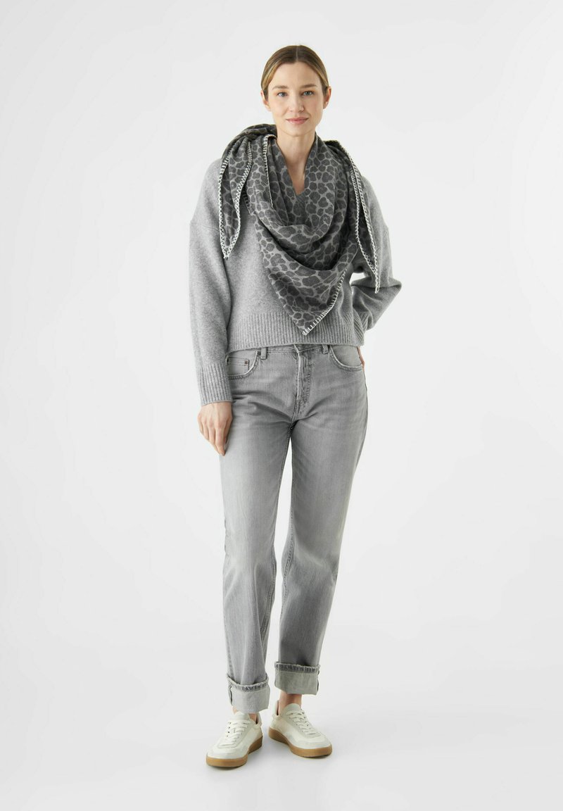 Femme portant un pull gris, une écharpe à imprimé léopard, un jean gris avec des ourlets, et des baskets blanches, se tenant devant un fond blanc.