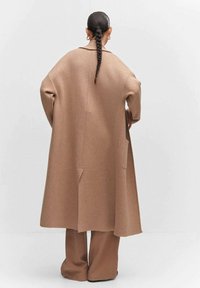 Long manteau taupe surdimensionné en tissu texturé semblable à de la laine, avec une fente à l'arrière et des poches latérales, associé à un pantalon large assorti.