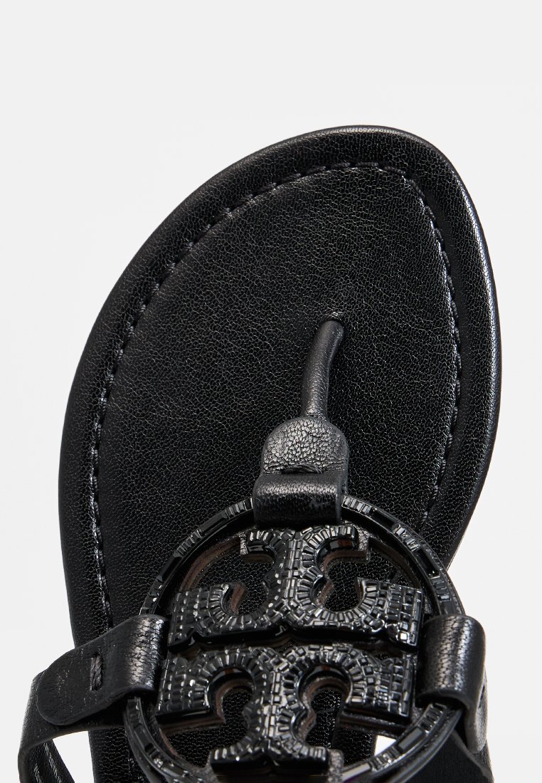 Schwarze Ledersandalette mit einem runden, strukturierten schwarzen Logo-Detail, schlanken Riemen und einer flachen Sohle mit glatter Oberfläche.