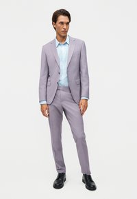 Άνδρας που στέκεται, φοράει ένα ανοιχτό μωβ κοστούμι με μπλε πουκάμισο και μαύρα παπούτσια πάνω σε απλό λευκό φόντο.