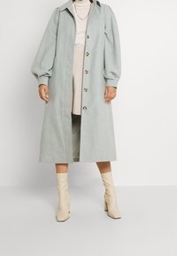 Manteau en mélange de laine bleu clair avec un devant à boutons, des manches bouffantes oversize et un col, associé à des bottes beiges jusqu'aux genoux.