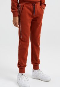 Rostfärgade svettbyxor i mjukt tyg, med sidofickor och vita accentränder. Matchas med vita träningsskor.