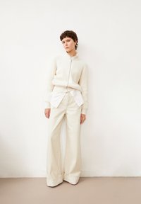 Crème ribbelgebreide zip-up trui over een wit shirt, gecombineerd met hoge taille, wijde lichtgekleurde broek. Eenvoudige schoenen maken de look compleet.
