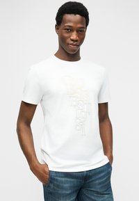 Άνδρας με κοντά σγουρά μαλλιά φοράει λευκό T-shirt GUESS με κείμενο και μπλε τζιν, στέκεται με τα χέρια στις τσέπες μπροστά σε μια απλή φόντο.